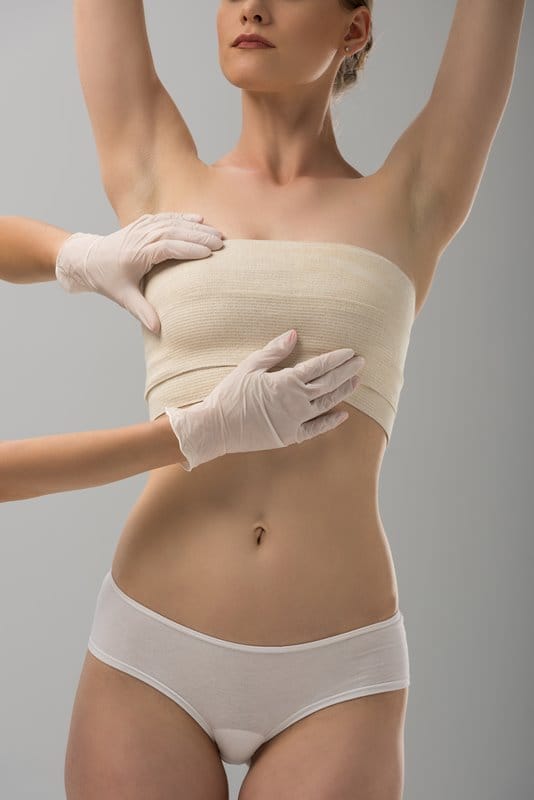 partial view of plastic surgeon in latex gloves and patient in breast bandage isolated on grey