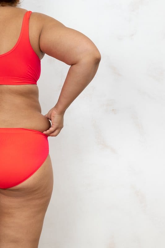 Photo of half of female cellulite body with back, free copy space, white background. Bare woman in red underwear pinch fat folds by hands. Overweight people, weight loss, self acceptance
